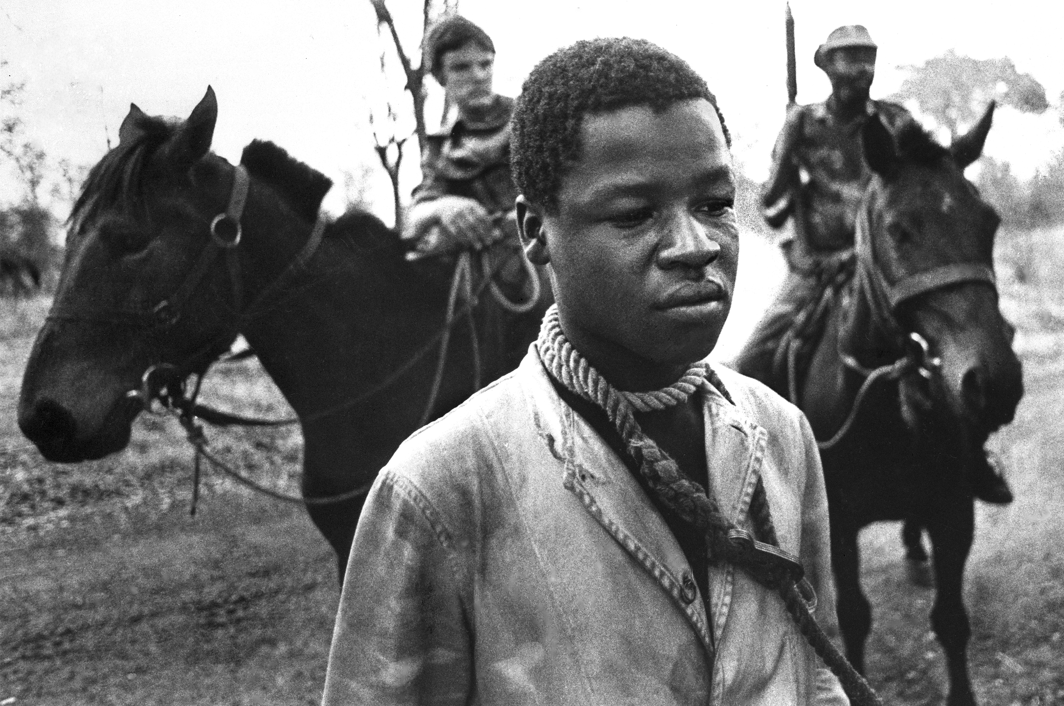 A Rhodesian prisoner stands with a rope tied around his neck to prevent escape, placed there by Rhodesian cavalrymen, background, who detained him for questioning in Lupane, southern Rhodesia, in September 1977. Rhodesia's white-dominated government was countering black guerrilla activities with militant armed forces. (AP Photo/J. Ross Baughman)